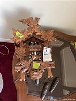Front view of carved wood cuckoo clock on glass table with pinecone weights and pendulum visible