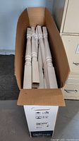 Photo showing white resin balusters packed in a tall cardboard box, viewed from above.