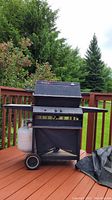 Front view of the black BroilKing Regal gas barbecue with propane tank under the shelf on the left side, placed outdoors on a wooden deck with greenery background.