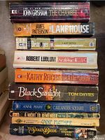 Stack of nine novels showing spines with titles and authors, including John Grisham's 'The Chamber', James Patterson's 'The Lake House', Robert Ludlum's 'Apocalypse Watch', and others.