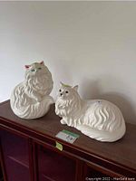 Two white porcelain cat figurines on a dark wooden surface against a white wall. One is larger and sitting upright, the other is smaller and lying down.
