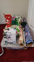 Wide view showing plastic tote with assorted Christmas wrapping paper rolls, gift boxes, green tree decoration, red bucket with snowman, and other holiday items