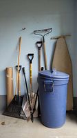 Blue Rubbermaid Roughneck 33 gallon garbage bin with locking lid and wheels, leaning against a garage wall with various garden tools and an ironing board.