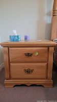 Front view of one oak color night stand showing two drawers with brass colored handles, top holds tissue box and purple candle.