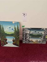 Two unframed landscape oil on board paintings displayed side by side on the floor against a wall, one with waterfall scenery, the other a mountain scene with deer.