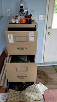 Beige filing cabinet with drawers partially open showing additional supplies inside and some items stacked on top such as tape, light bulbs.