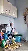 Wide angle photo showing the collection of various garden and cleaning supplies including watering can, cleaning liquids, compost bucket, blue bucket, paint supplies and broom.