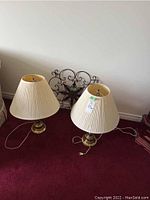Two metal table lamps with cream pleated lampshades showing wear and a black metal wall art piece behind them.