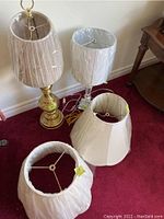 Two table lamps and four lamp shades grouped on carpeted floor, showing brass and glass bases and shade variety