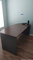 Front angled view of Bush office desk showing dark wood top, curved front edge, and black side panels on wood flooring.