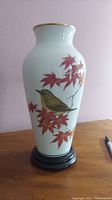 Front view of porcelain vase showing yellow-green bird and red maple leaves with gold rim and dark wood stand