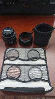 Photo shows Olympus Zuiko 50mm lens with lens hood, black drawstring pouch, and a folded filter case with four filters inside.