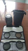 Olympus 14-45mm camera lens, black padded carrying pouch, and camera lens filters stored in a fabric pouch with mesh pockets on a wooden surface.
