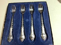 Four ornate stainless steel serving forks laid inside a blue velvet-lined box, photo showing overall set condition and presentation.