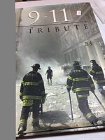 Front cover of the 9-11 Tribute book showing firefighters in F.D.N.Y. gear walking through rubble in a smoky urban disaster scene.