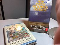 Photo showing two hardcover books: 'At the Cottage' by Charles Gordon with illustrated dust jacket showing a cottage dock scene, and 'How I Spent My Summer Holidays' by W.O. Mitchell with a dark cloudy sky dust jacket.