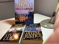 Three paperback books by David Baldacci standing and laying on a table, titles: Hell's Corner, Stone Cold, Divine Justice, with visible cover images and text.