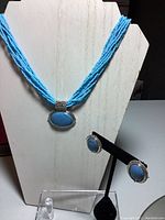 Photo of the blue stone necklace displayed on a white stand alongside matching oval blue stone earrings on a black stand, showing the multi-strand seed bead necklace and silver-tone settings.