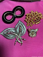 Four costume pins including cat face with moving eyes and pink ear, gold-toned basket of flowers, black figure 8 or mask pin, and silver-toned leaves with some missing rhinestones, all placed on a pink cloth background.