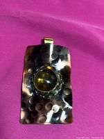 Photo of rectangular hammered copper pendant with central clear green circular glass or stone on magenta background.