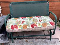 Green metal frame garden glider with mesh back and seat, floral-patterned cushion placed on the seat outdoors on a porch with wooden siding background.