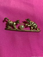 Gold toned cat-themed brooch with two cats and a pearl between them on a textured horizontal bar, photographed against pink background.