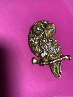 Gold-toned owl pin with rhinestone accents on a bright pink background
