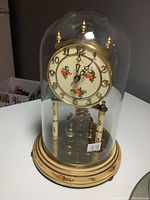 Front and slightly angled view of Kieninger & Bergfell mantle clock showing floral decorated face, gold-tone numerals and rotating pendulum under glass dome on floral base.