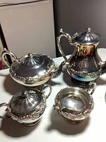 Four-piece silver plated tea and coffee service set displayed together showing teapot, coffee pot, creamer, and sugar bowl with ornate floral edging and shiny finish.