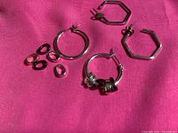 Two pairs of sterling silver hoop earrings displayed on a pink fabric background. One pair is round with three small oval charms, the other pair is hexagon shaped.