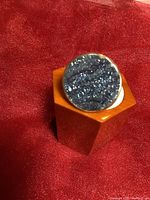 Front view of the sterling silver adjustable ring with round midnight blue druse stone top, placed on a hexagonal orange stand against a red background.