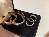 Photo showing two pairs of sterling silver hoop earrings placed in a small jewelry box, one pair smaller with intricate pattern and safety clasp, the other larger with hammered finish.