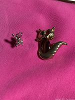 Two sterling silver items on pink fabric background: a small mouse pendant on the left, and a larger cat pin on the right with amber body and colored gemstone eyes.