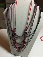 Front view of trio of seed bead necklaces on white mannequin. Necklaces include black 3-strand seed bead necklace, red single-strand seed bead necklace with larger stones, and purple 3-strand seed bead necklace with crystal accents.