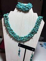 Turquoise stone necklace draped on white display stand with matching bracelet at top and earrings hanging on black stand.