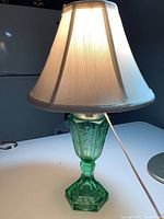 Photo showing front and side view of vintage green glass table lamp with off white shade lit from bulb inside.