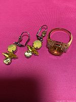 Photo showing one gold toned ring with large yellow and clear crystals, and one earring showing yellow and green crystals with safety clasp on pink background.