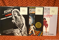 Photo showing three vinyl soundtracks with numbered stickers 141 on the top right corner of each album sleeve. Titles include The Rose, Saturday Night Fever, and Lady in Red.