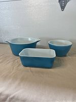 Three blue Pyrex kitchenware items displayed in a row on a beige cloth background, showing the shapes and colors of the Fridgie dish, bowl, and casserole dish.