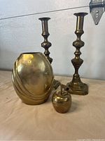 Photo showing all four brass items: vase, apple bell, and two candlestick holders, placed on a cloth surface with a neutral background.