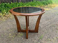 Front view of round walnut side table with smoked glass top sitting outdoors on gravel, with plants and wooden fence in the background showing wood grain and glass condition.