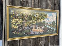 Needlepoint artwork in gold ornate frame, showing the full piece including the large tree, people, dogs, river, and sky.