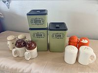 Wide shot of 3 vintage metal canisters labeled Coffee, Sugar, and Flour with black lids, and 4 pairs of salt and pepper shakers of various styles including avocado, orange, white ceramic with handles and brown tops.