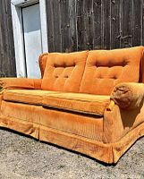 Front angled view of the vintage orange corduroy loveseat showing tufted back cushions, pleated skirt base, and rolled arms with bolster pillows.