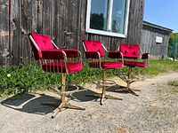 Three vintage red velvet bar stools arranged side by side outdoors against a wooden wall with clear daylight.