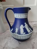 Blue Wedgwood jasperware pitcher with white relief classical figures, handle with leaf motif details.