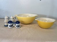 Front view of two yellow Pyrex mixing bowls with white interior alongside four salt and pepper shakers, two cream and blue, two white and blue with dog illustrations.
