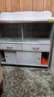Front view of vintage arborite cabinet showing two glass sliding doors with etched design, two drawers and two sliding doors below, with a small orange funnel visible on bottom shelf.