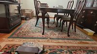 Wide angle photo showing the carpet placed under a dining table set in a room, carpet has fringe along edges and floral patterns.