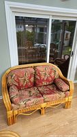 Full view of rattan couch with two floral cushions and an armrest pillow on hardwood floor in front of sliding glass door.